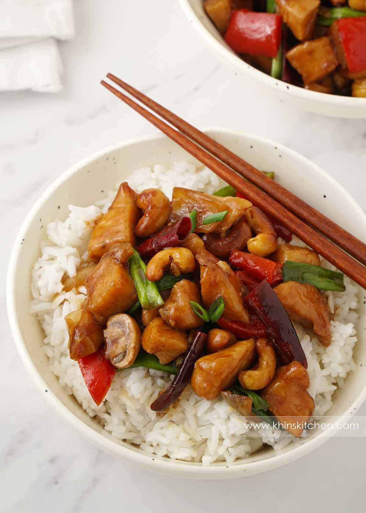 A white bowl containing chicken stir fries, cashew nuts, mushrooms, chillies and white rice.