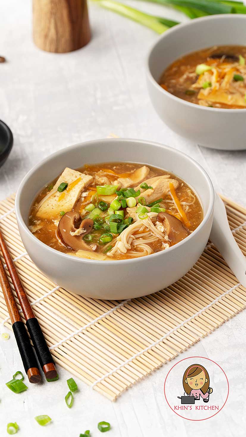 A grey bowl containing thick glossy hot and sour soup with mushrooms, tofu, shredded carrot, egg ribbons, and spring onions. 