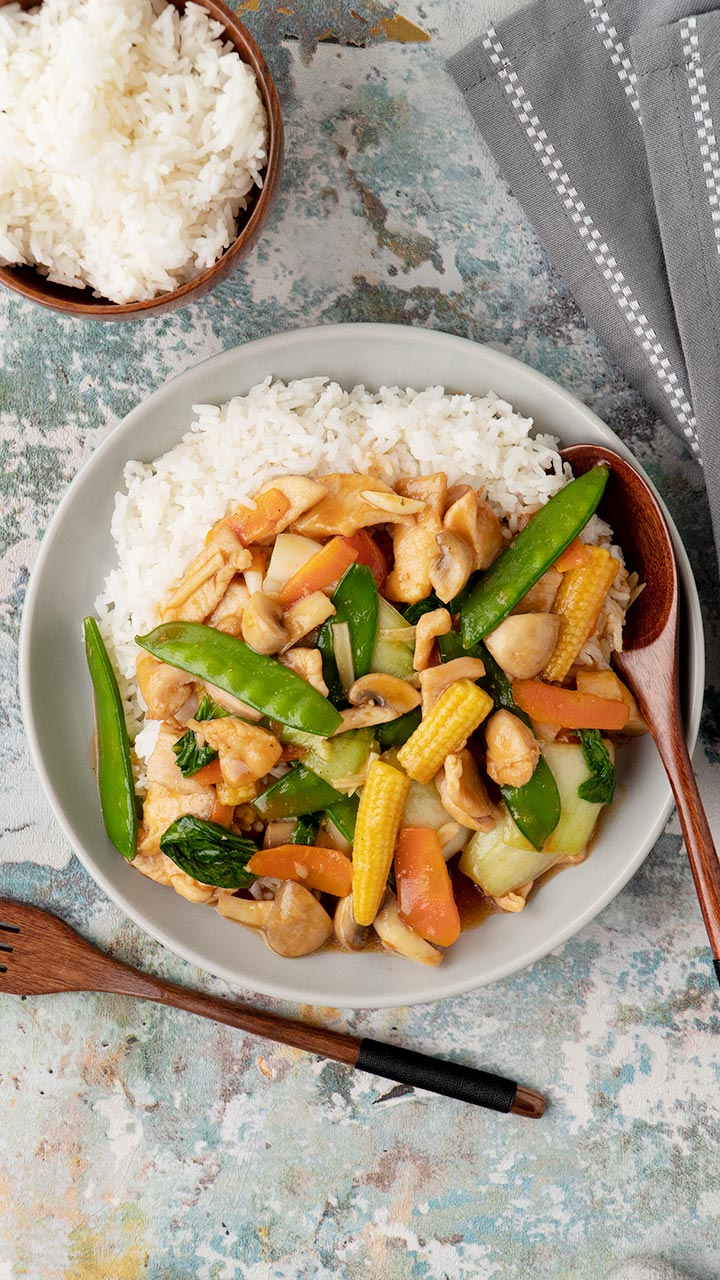 A grey plate containing rice topped with Chinese chicken stir fry with mushroom, vegetables, and glossy sauce. 