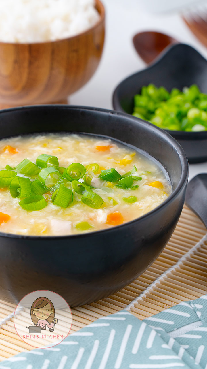 A black bowl containing soup with egg drops, carrots, chicken, sweetcorn and chopped green onions. 