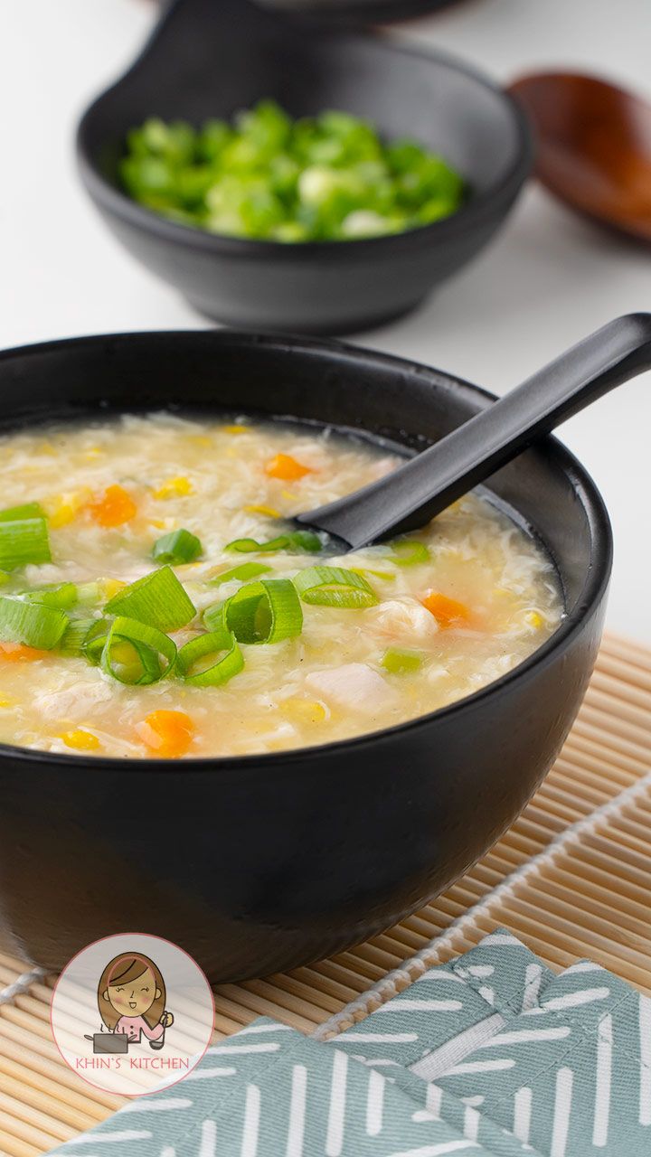 A black bowl containing soup with egg drops, chicken, carrot, sweetcorn, and spring onions. 