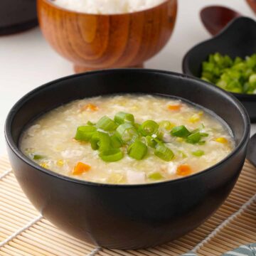 A black bowl full of Chinese style chicken and corn soup, topped with spring onion in the foreground and a small wooden bowl of white rice in the background.