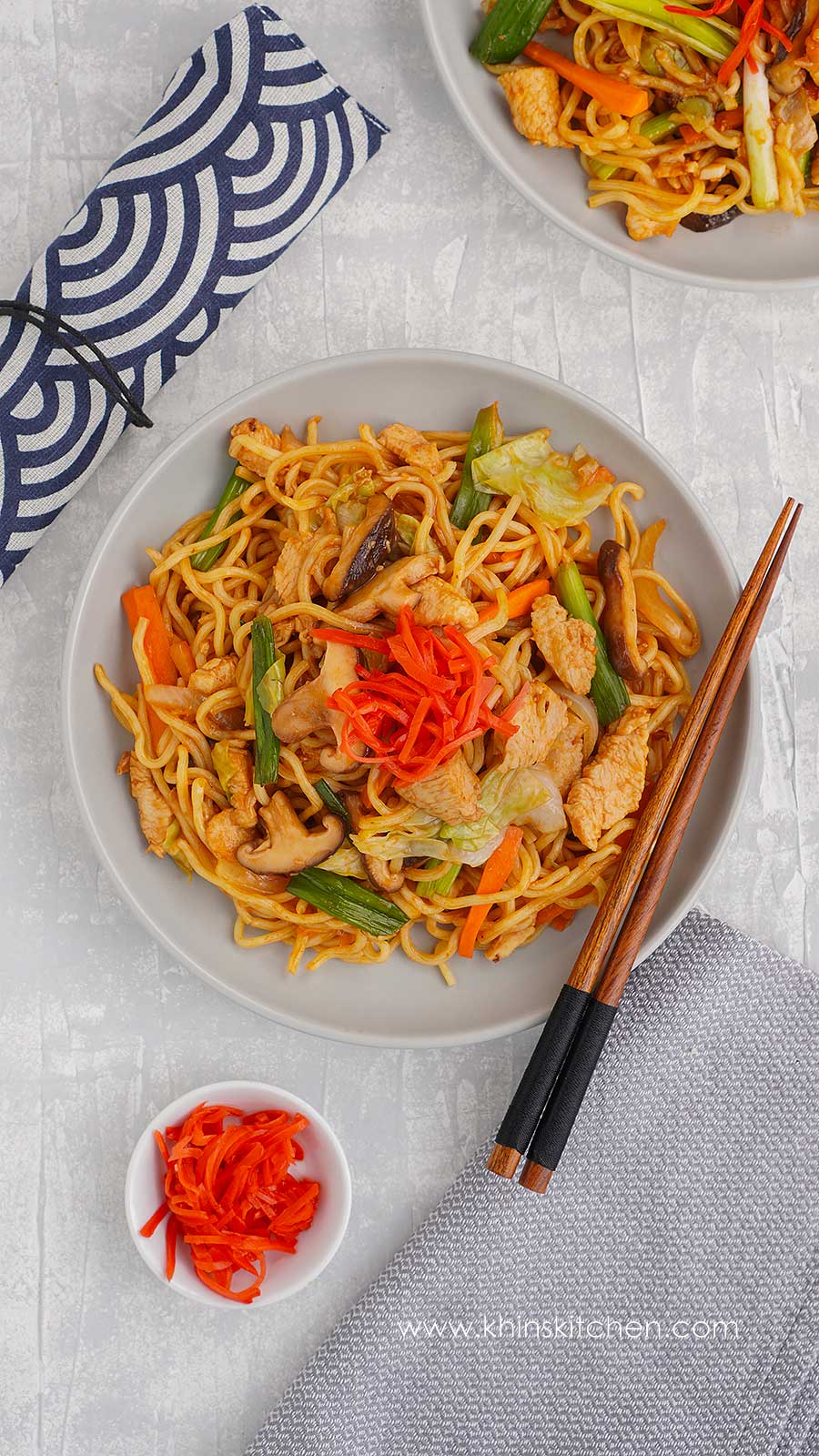 A grey plate containing Japanese-style stir-fried noodles with chicken, shiitake mushroom, colourful vegetables, and ginger pickle on top. 