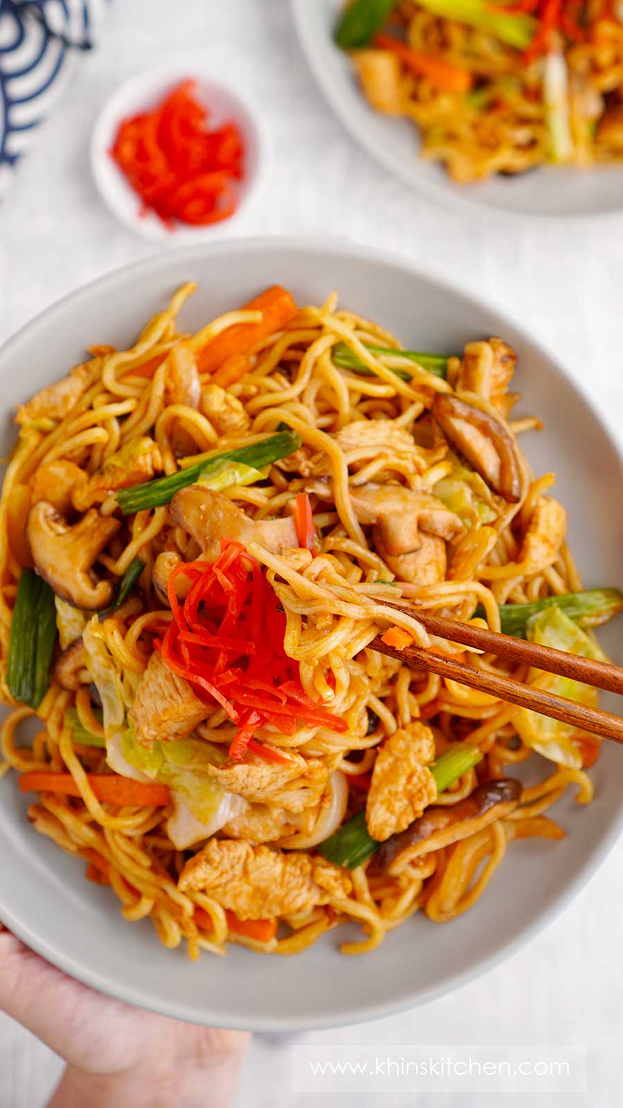 An image showing a wooden chopstick holding noodles and a grey plate containing Japanese fried noodles with vegetables and ginger pickle. 