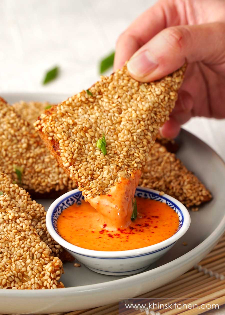 A hand holding a triangle shape sesame shrimp toast dipped in a spicy mayonnaise.