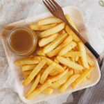 A wooden plate with chips and curry sauce in a jar and wooden spoon on the plate.