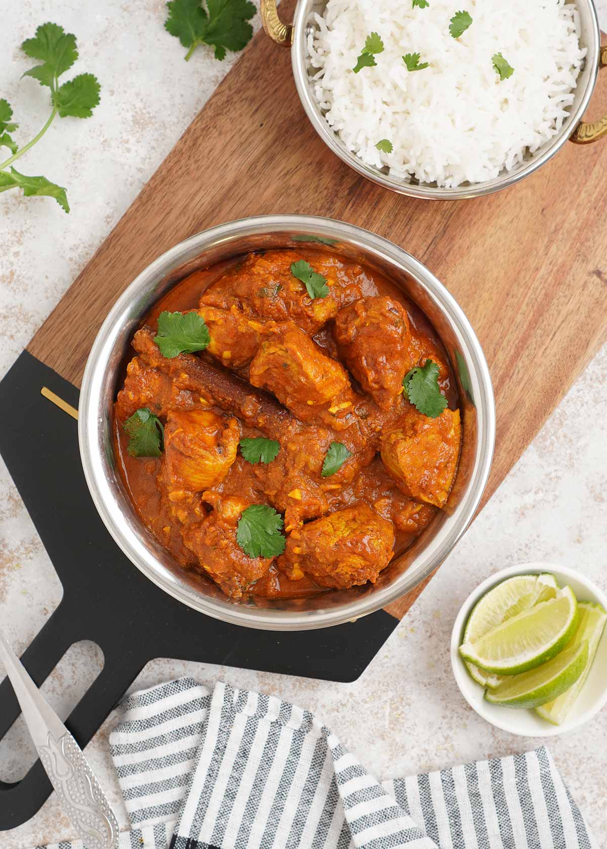 chicken curry in a steel pot with a small bowl of white rice on the wooden serving board and a small bowl of lemon wedges for garnish.