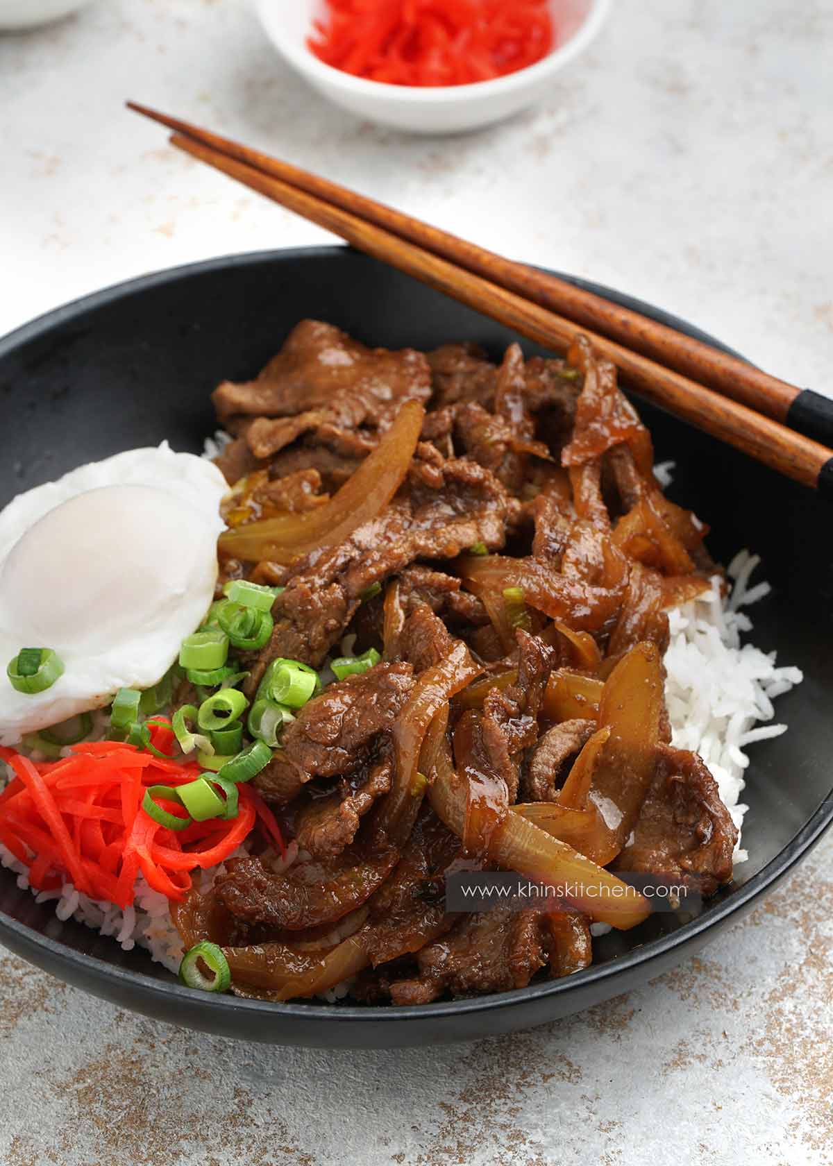 A bowl of japanese style beef stir fry with egg on the side and pickle.