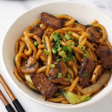 A white bowl containing stir fry udon noodles, stir fry beef slices and finely chopped spring onions