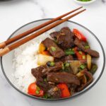 a white bowl containing stir fried beef, stir fried vegetable and sprinkle of spring onions.