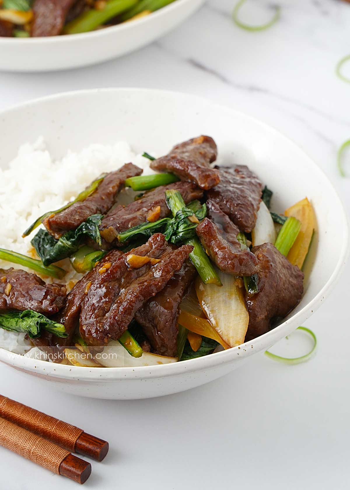 A white bowl containing plain rice, stir fry beef and green vegetables. Wooden chopstick on the left corner. 