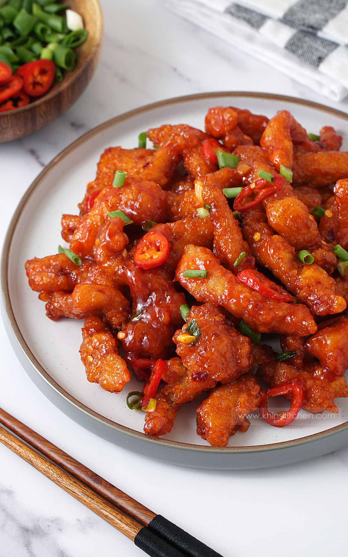 A white plate containing, spicy red fried chicken strips with chopped green onions and chillies. 