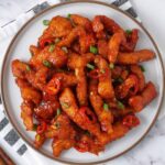 A white plate containing, spicy fried chicken strips, topped with spring onion and sliced chillies.