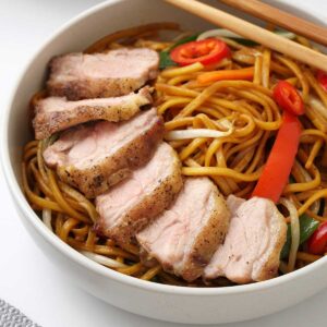 A white bowl containing stir fried noodles topped with roasted sliced duck.