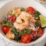A mixture of prawn salad served on the white bowl consisting of cooked prawn, glass noodles, coriander, tomatoes and lime.