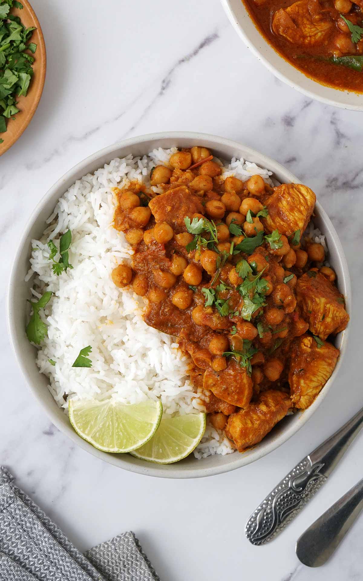 A grey bowl containing white rice, chicken and chickpea in curry sauce with lime wedges on the side. 