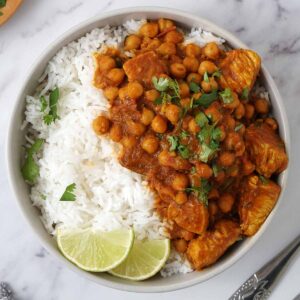 A grey bowl containing chicken curry with chickpea and topped with coriander and lime wedges.