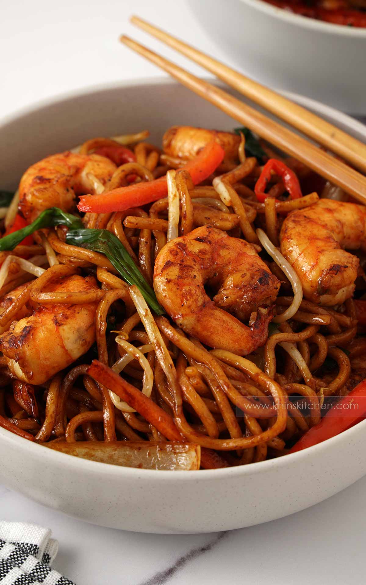 A grey bowl containing brown noodles, prawns and vegetable stir fry. Wooden chopstick on the side. 