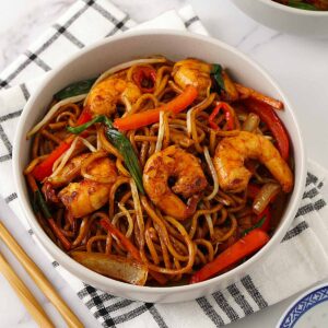 A white bowl containing stir fried noodles, stir fried vegetables and king prawns on the white tea towel.