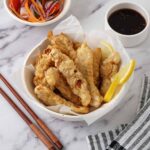 A white bowl containing sliced fried chicken, lemon wedges and a side of small dipping sauce.