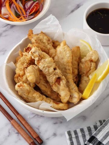 A white bowl containing sliced fried chicken, lemon wedges and a side of small dipping sauce.