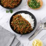 A black bowl containing lamb mince curry on top the white rice with a small bowl lemon wedges, a small plate of sliced green chilies and steel cutlery.
