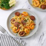 A grey plate containing pasta, sliced chorizo and prawn, sprinkle with parsley and grated cheese.