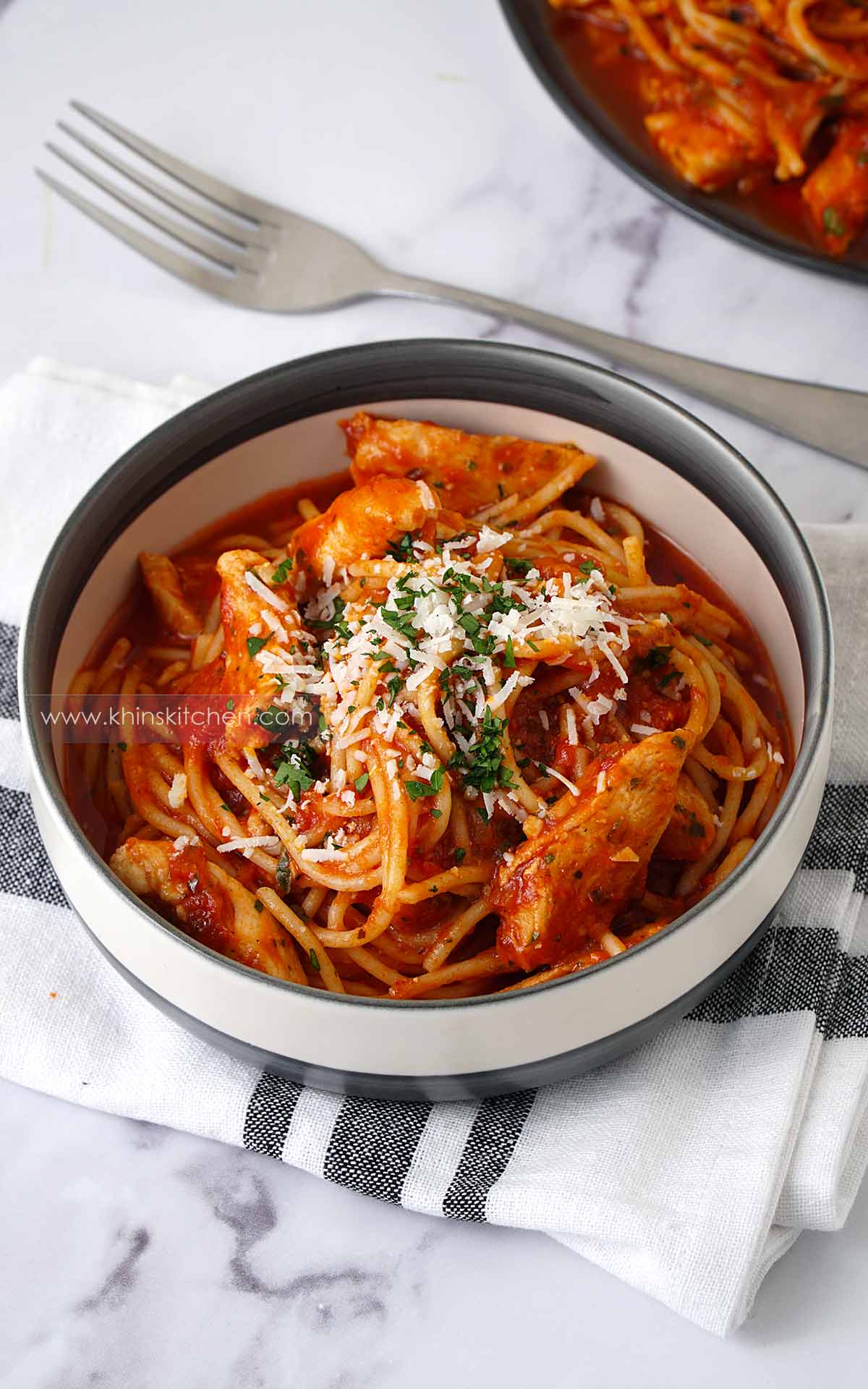 A white & grey bowl containing spaghetti, chicken slices and red tomato sauce. Topped with grated cheese and finely chopped parsley. 