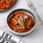 A white bowl containing pasta and chicken with tomatoes sauce and sprinkle with cheese and parsley.