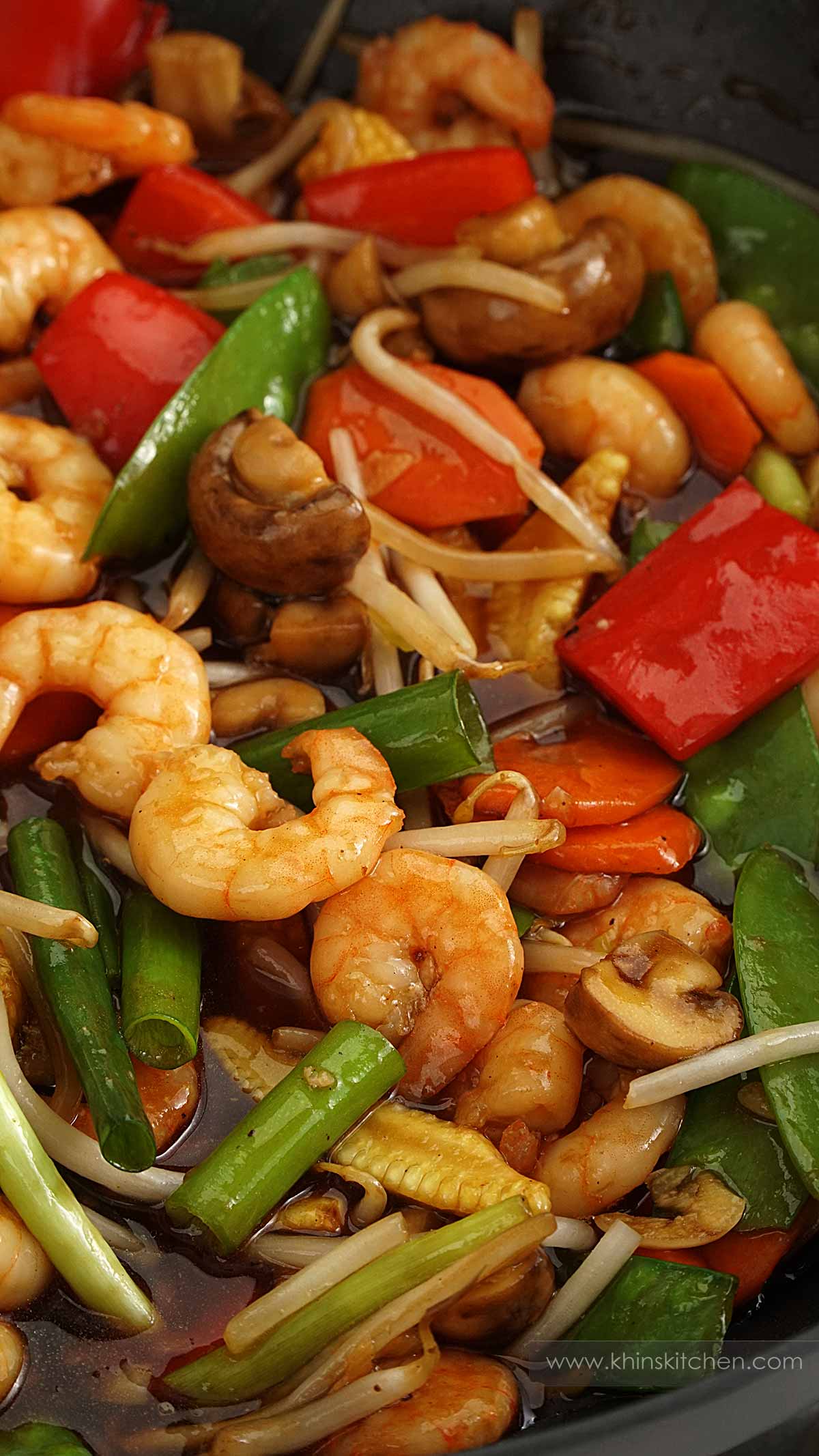 A large wok containing stir fry, king prawns, bean sprouts, bell peppers, mushroom, mangetout, carrot and baby corn with a glossy brown sauce. 