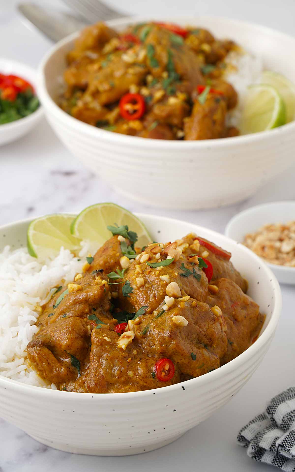 A white bowl containing peanut butter chicken curry, topped with coriander, red chillies, and crushed peanuts. 