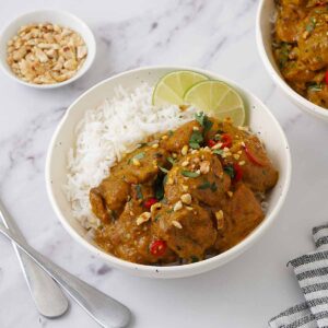 A white bowl filled with chicken curry on top of the white rice, sprinkle with crushed peanuts, coriander and two slices of lime.