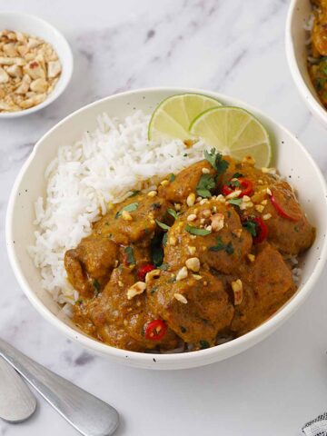 A white bowl filled with chicken curry on top of the white rice, sprinkle with crushed peanuts, coriander and two slices of lime.