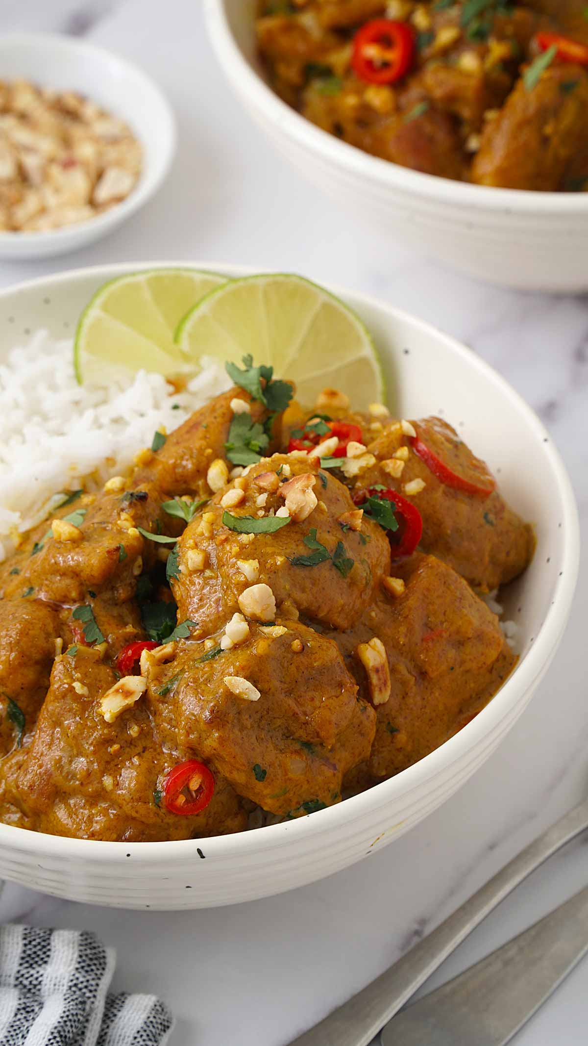 A white bowl containing Chicken Curry, crushed peanut, white rice and lime wedges.