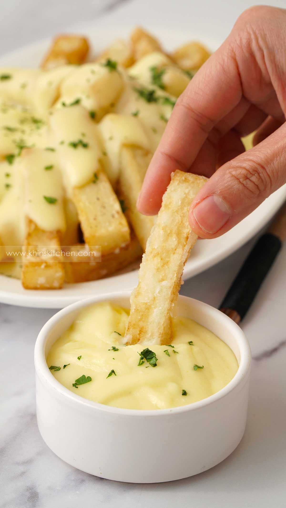 Fried golden chip is held by two fingers and half dipped in a cheese sauce with chopped parsley. 