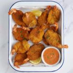 A white tray containing golden brown butterfly king prawns and tomato dipping sauce on the side.