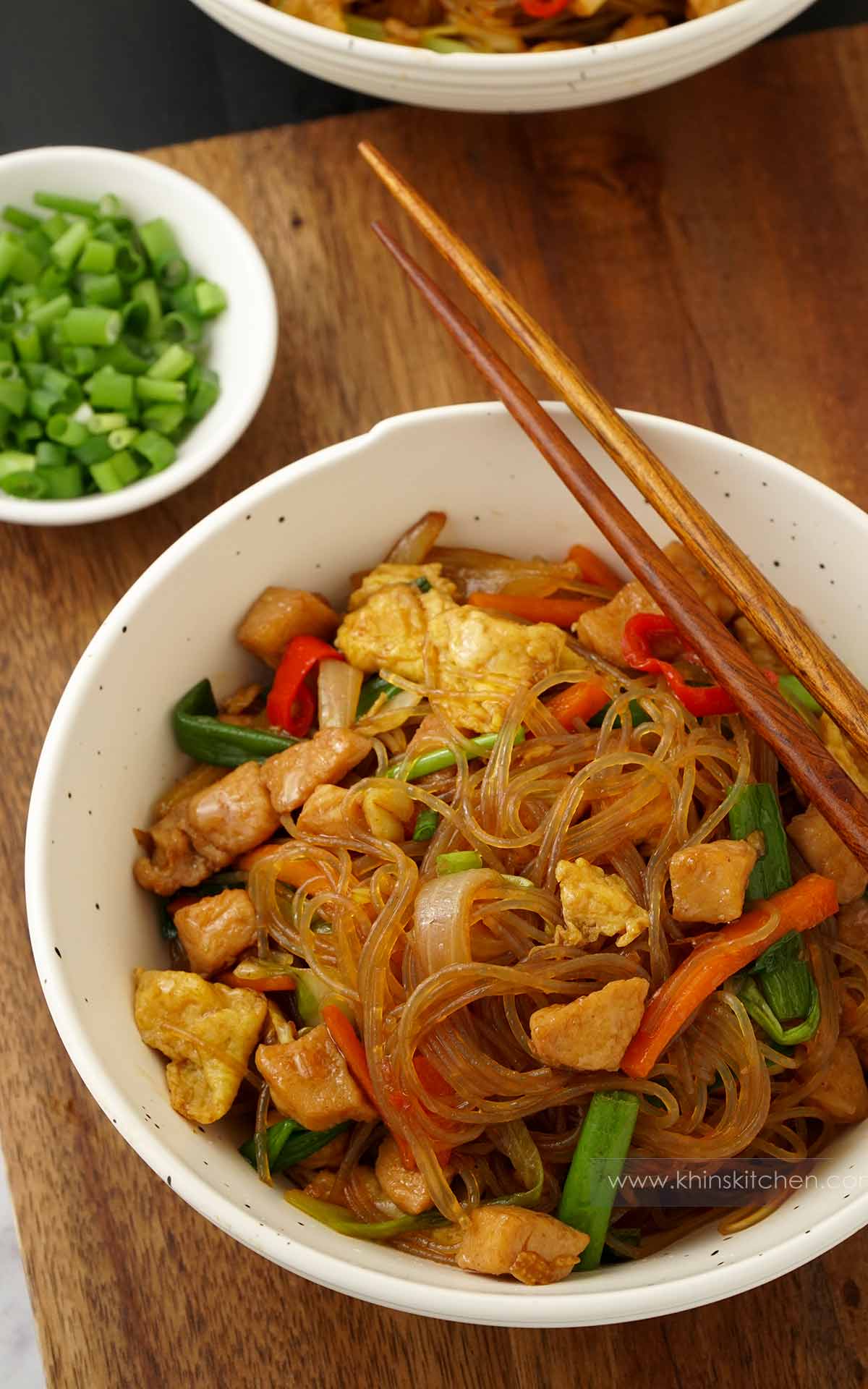 A white bowl containing fried glass noodles with chicken, egg and vegetables. Wooden chopstick on the side. 