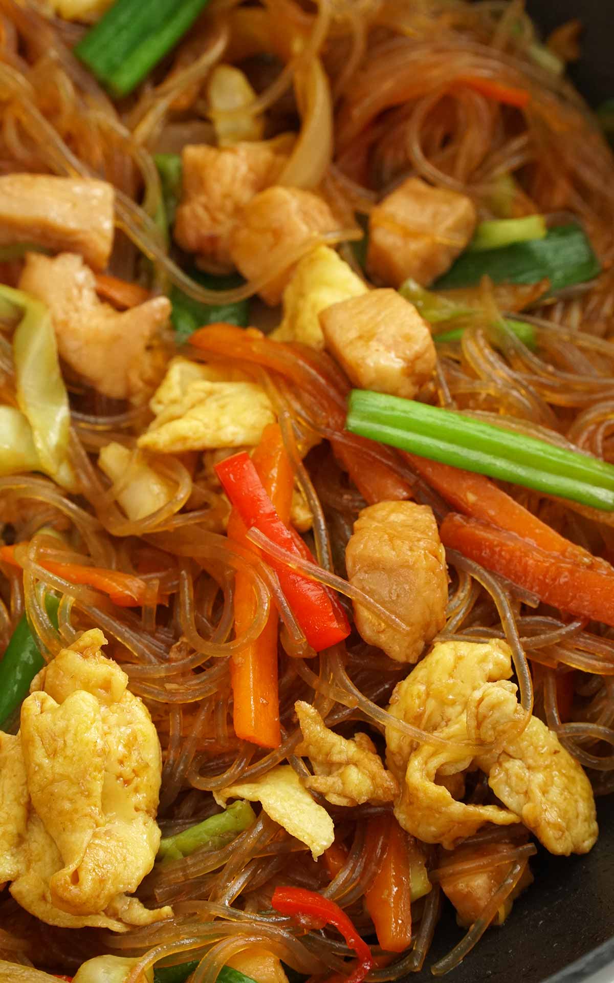 A large wok containing stir fried glass noodles with carrot, spring onions, pepper, chicken and eggs. 