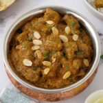 A copper serving bowl containing lamb curry with creamy sauce and almond flakes.