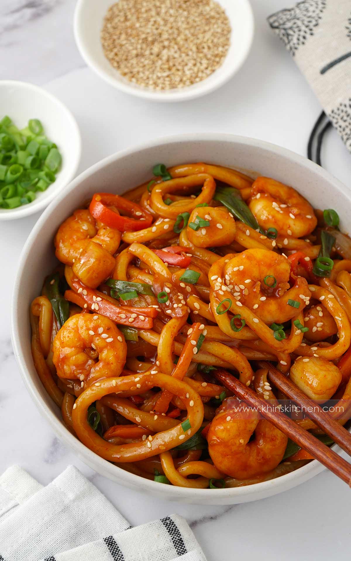 A white bowl containing stir fried udon noodles, prawns, squid rings, scallops, and red chilli slices. Spring onions and sesame seeds on the top.