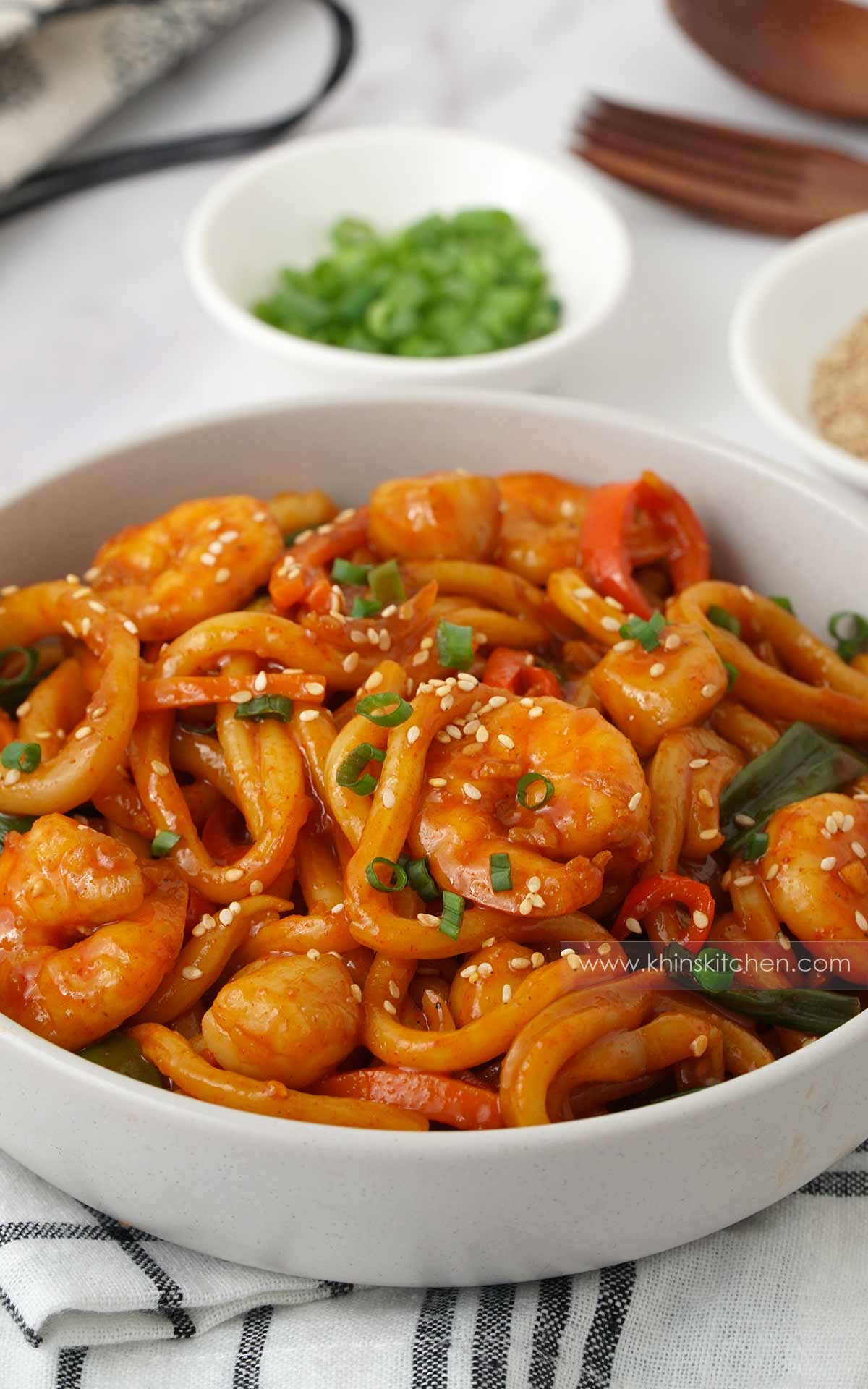 A white bowl containing thick round noodles with shrimp, scallops, squids, spring onions and sesame seeds with red spicy sauce. 