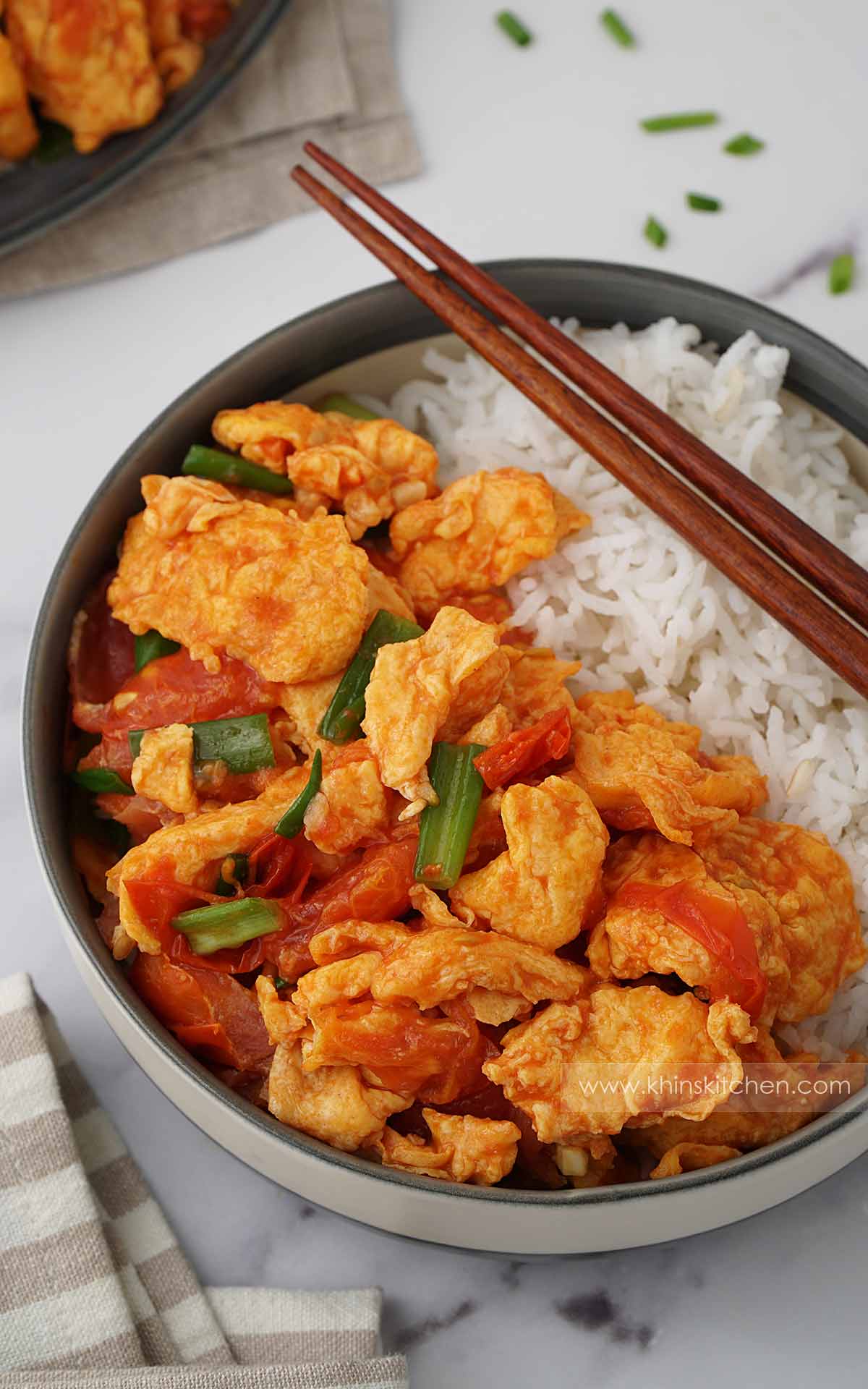 A grey bowl containing stir fried tomatoes, egg and spring onions with white rice. Wooden chopstick on the side. 