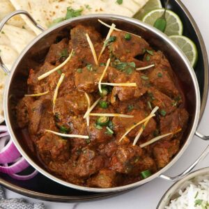 Steel serving bowl containing lamb curry with fresh ginger strips, green chillies and coriander. Onion slices, lime wedges and naan on the side.