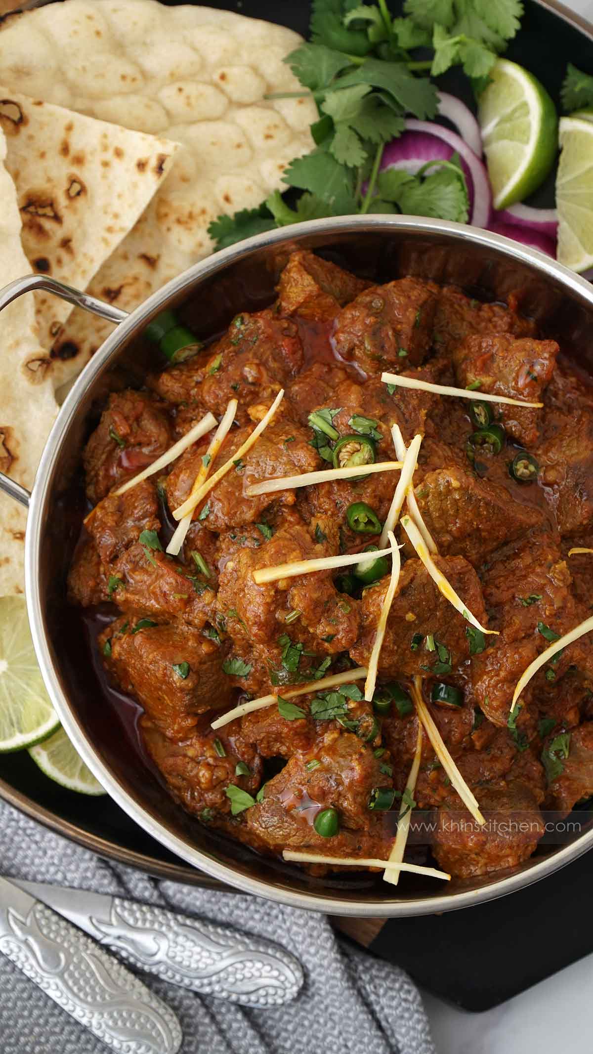 A metal bowl containing lamb curry in karahi sauce, garnish with ginger strips, green chillies and coriander. 