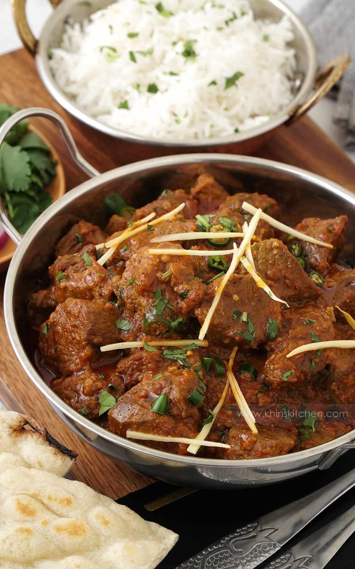 A steel serving bowl containing lamb curry. A rice bowl and naan on the side. 