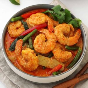 A white bowl containing, prawns and vegetable with creamy red curry sauce. Coriander on the side.