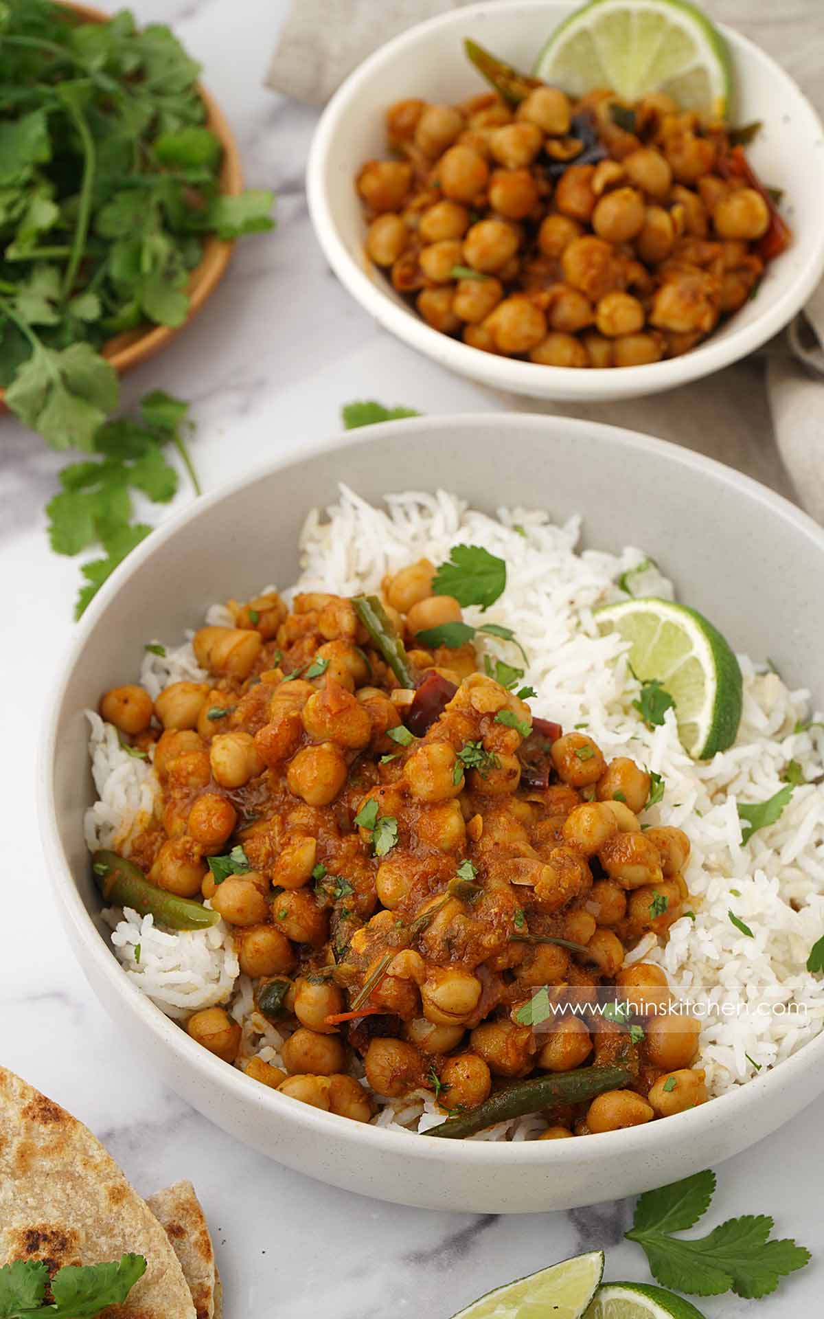 A grey bowl containing chickpea curry and basmati rice. Lime wedges and coriander on the side. 