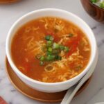 A white bowl containing egg drop soup with tomatoes and spring onions.