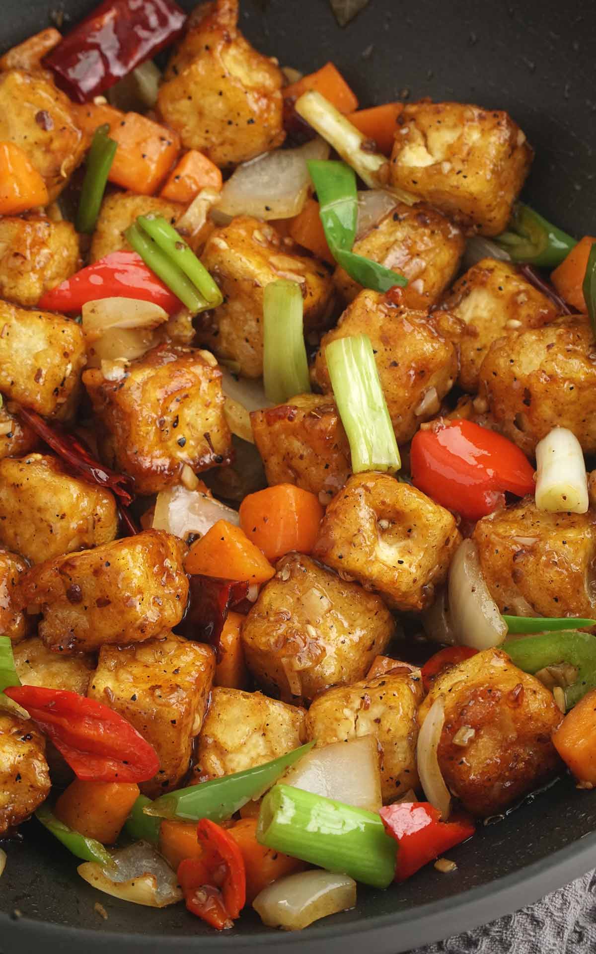 A black wok containing fried tofu cubes coated in sauce together with dried chillies, Szechuan peppercorns, and vegetables.