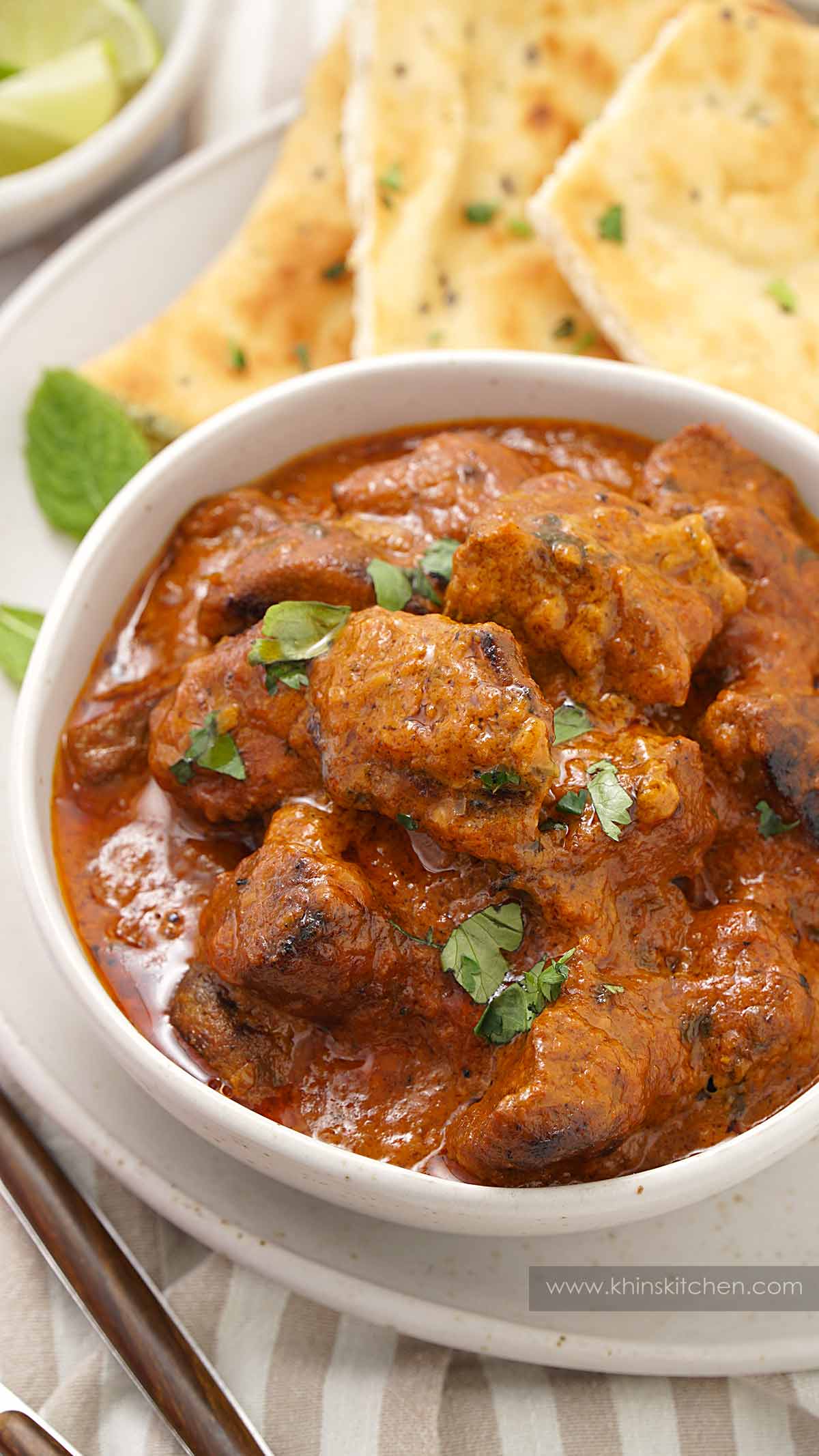 A white ceramic bowl containing, lamb tikka pieces with creamy masala sauce. Naan bread on the side.